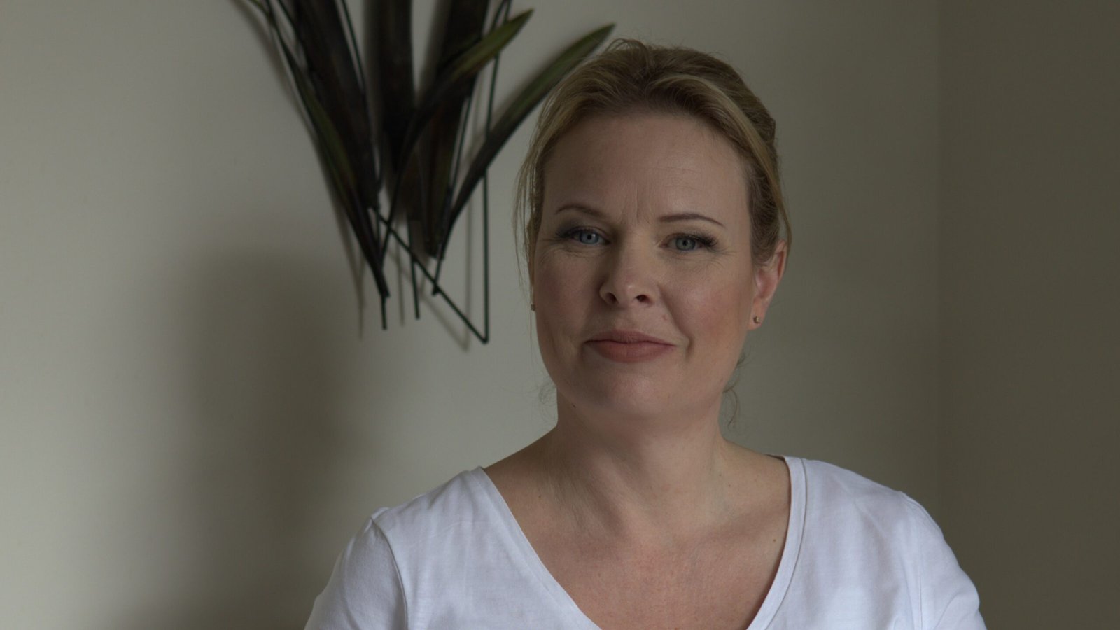 Natasha smiling warmly at the camera, wearing a white V-neck t-shirt, standing in front of a pale wall with dark green decorative leaves.